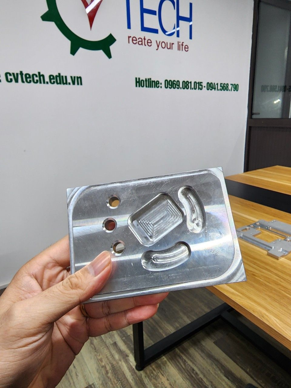 Chi tiết nhôm phay cầm trên tay trước phông logo CVTECH - gia công cnc 4 trục