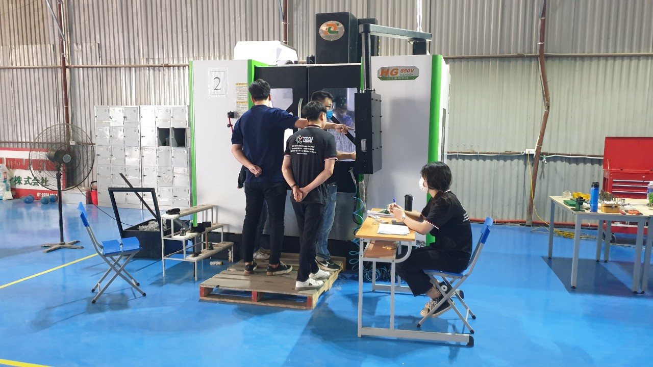 Nhóm học viên đứng trên bục thao tác máy phay CNC, giảng viên ngồi bàn bên cạnh - tính tốc độ cắt khi phay cnc